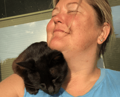 Heidi smiling peacefully with eyes closed while her black cat nuzzles her cheek, reflecting self-compassion and warmth