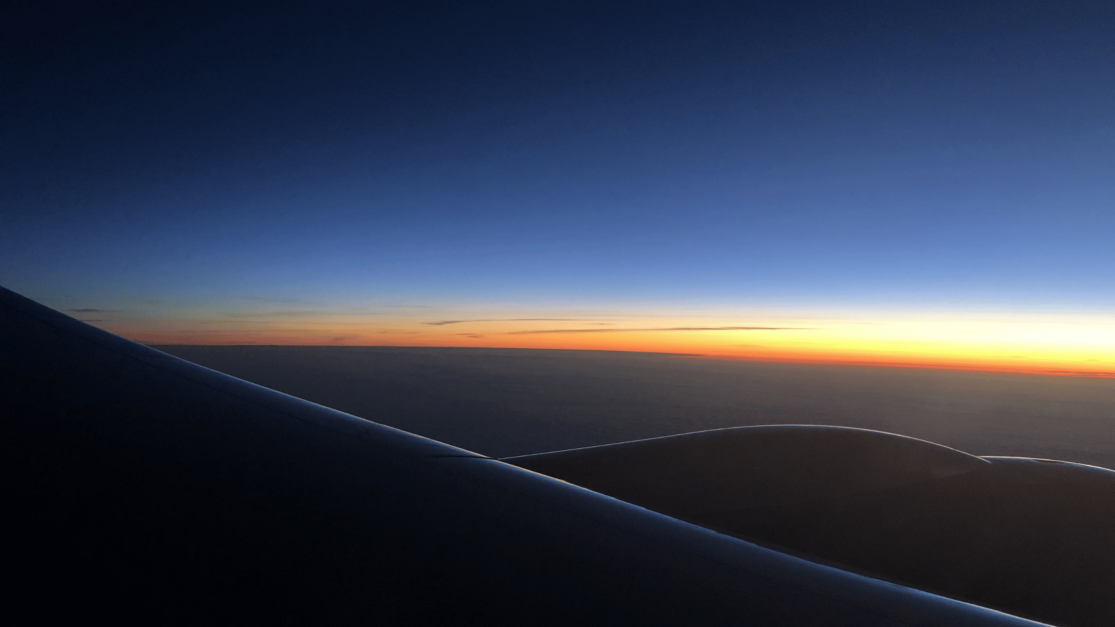A panoramic airplane window view at dawn, showing the dark curve of the wing against a vibrant horizon of deep blue and orange, representing the liminal space and maternal energy discussed in Heidi’s blog.