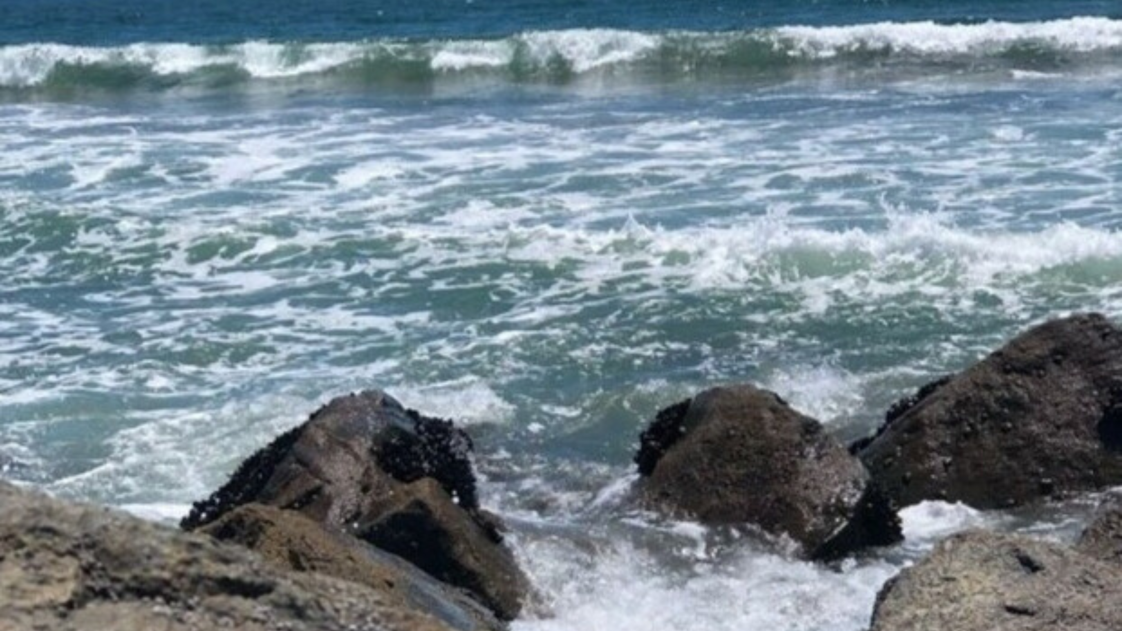 Waves crashing against rugged rocks under a clear blue sky, symbolizing the ebb and flow of emotions and the strength found in facing discomfort.